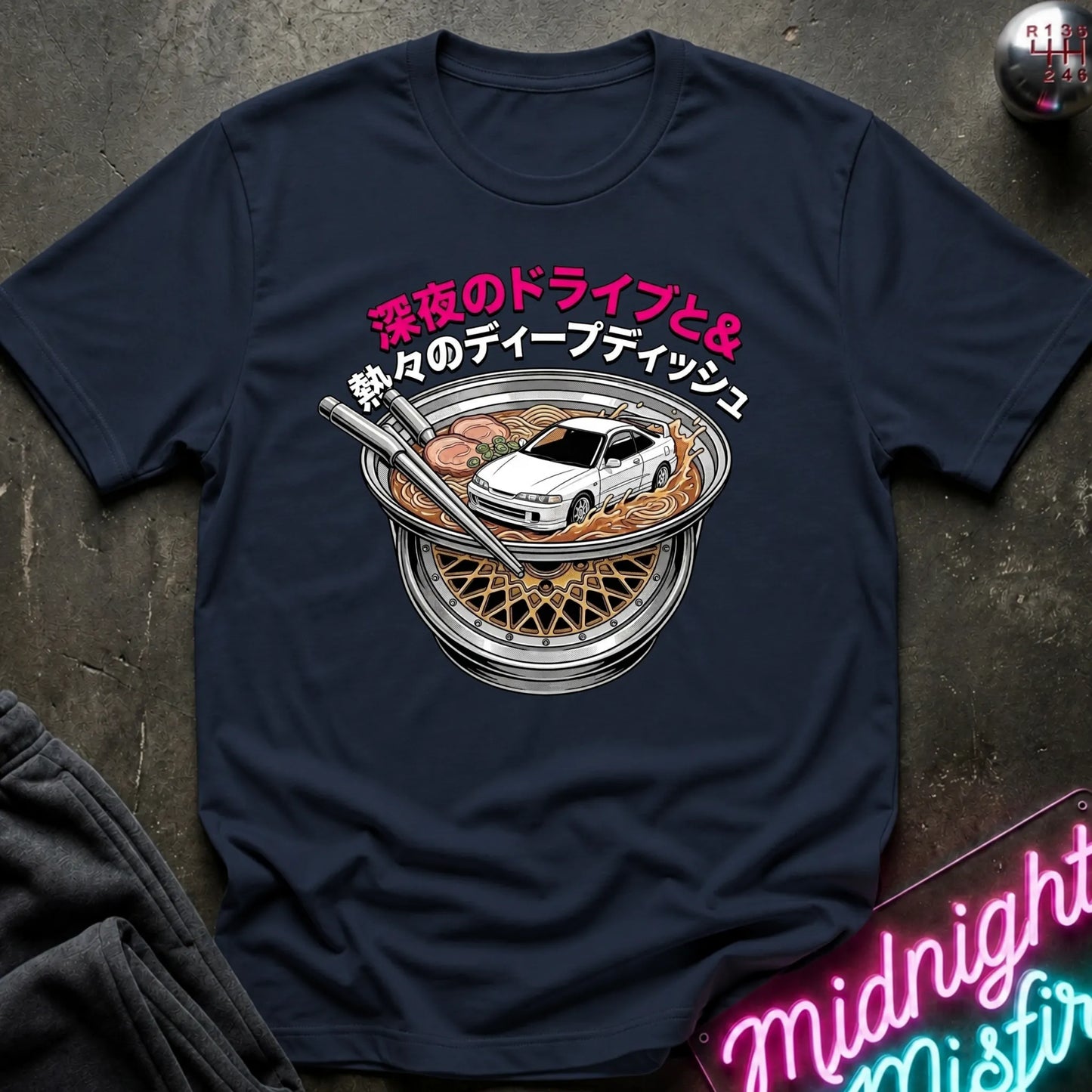 A navy blue graphic t-shirt laid flat on a dark textured surface. The shirt features a circular graphic of a white sports car inside a bowl of ramen with chopsticks, accompanied by pink and white Japanese text.