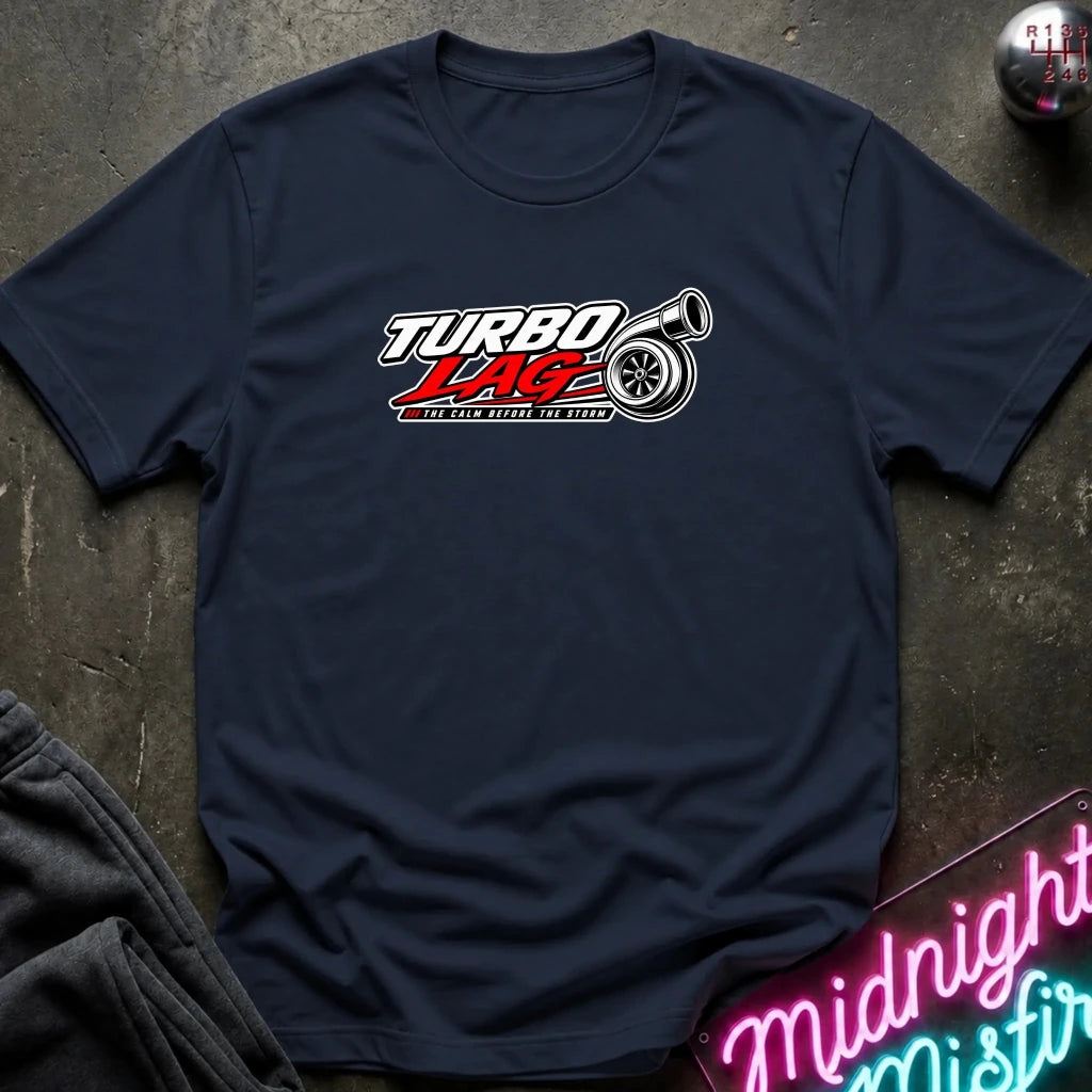 A navy blue short-sleeve t-shirt with a 'Turbo Lag' muscle car graphic on the chest, laid flat on a dark textured surface with a neon sign and dark pants in the background.