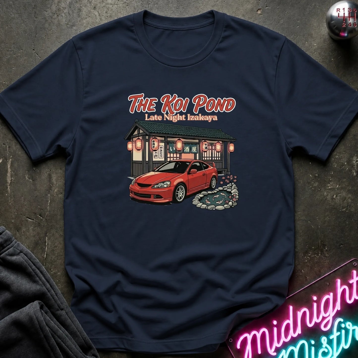 A navy blue short-sleeve graphic t-shirt laid flat on a dark textured surface. The graphic features the text 'THE KOI POND Late Night Hamburger' above an illustration of a red sports car parked in front of a restaurant with red lanterns and a koi pond.