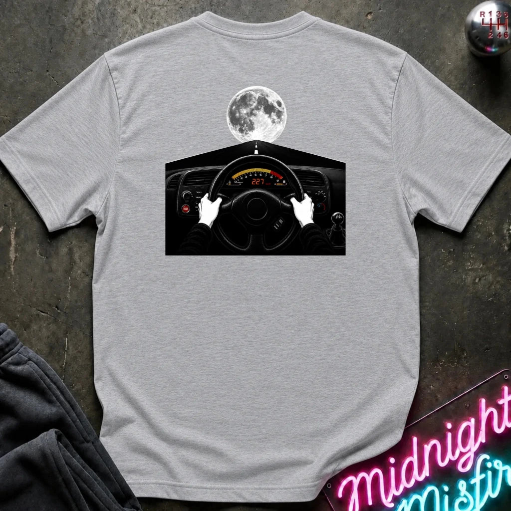 A heather gray t-shirt laid flat on a dark textured surface, featuring a graphic print of a driver's hands on a steering wheel with a full moon above. A neon sign and folded sweatpants are partially visible as props.