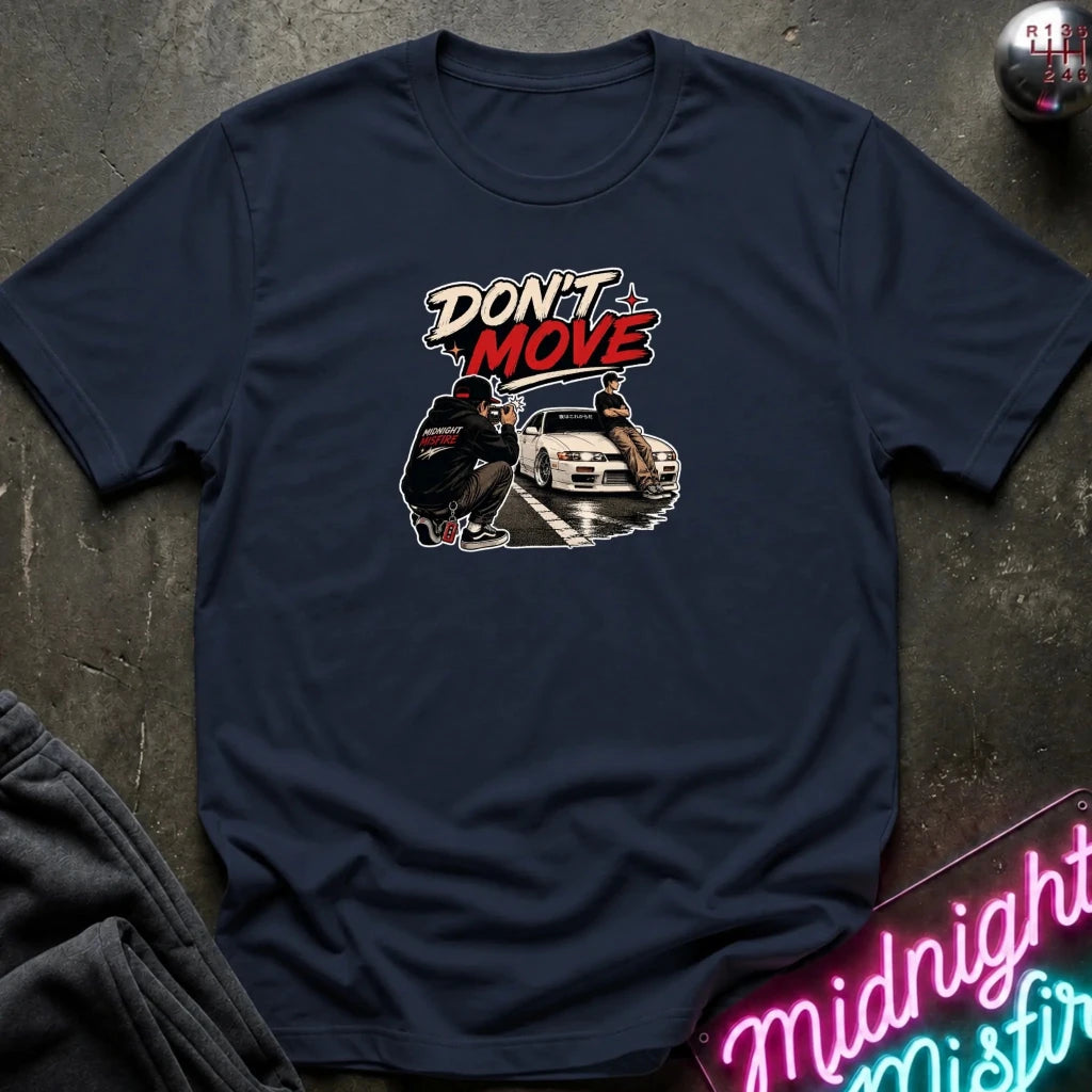 A navy blue short-sleeve t-shirt with a graphic print on the chest featuring the text 'DON'T MOVE' in white and red, above an illustration of a person and a white car. The shirt is laid flat on a dark textured surface with a neon sign and dark pants partially visible in the corners.