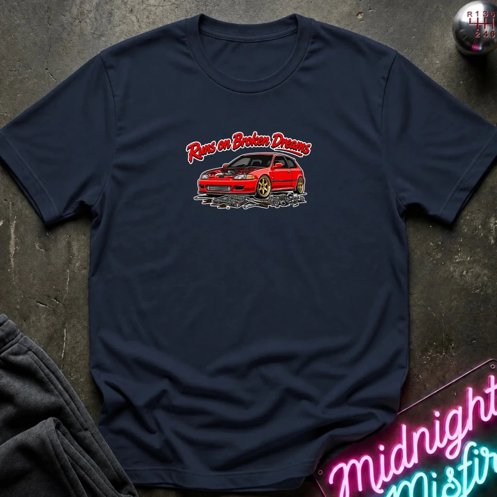 A navy blue crewneck t-shirt laid flat on a dark textured surface. The shirt features a graphic print of a red car with the text 'Run on Broken Dreams' in red and white script.