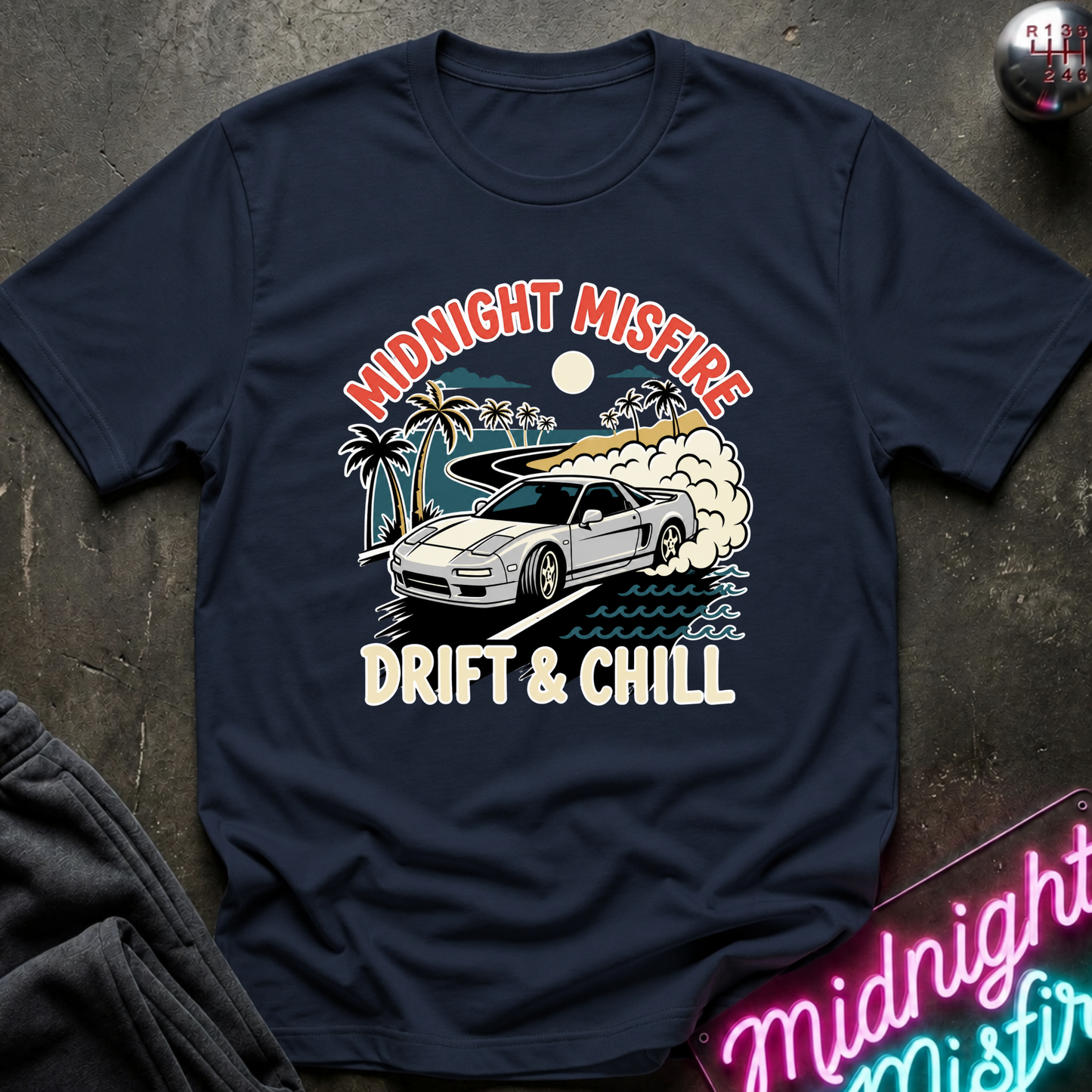 A navy blue graphic t-shirt laid flat on a dark textured surface. The shirt features a retro-style print of a white sports car drifting on a beach with palm trees, waves, and the text 'MIDNIGHT MISFIRE' and 'DRIFT & CHILL'.