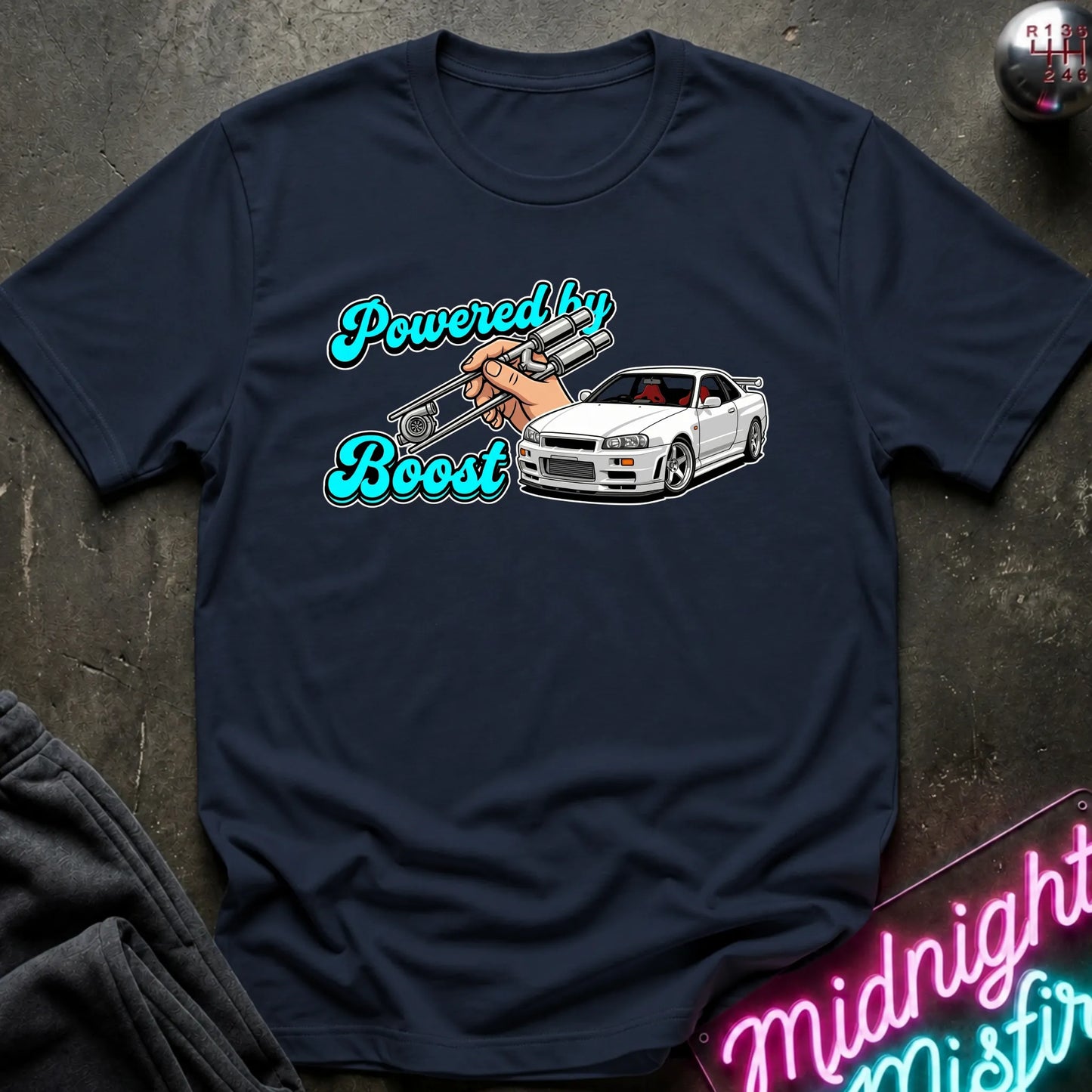 A navy blue graphic t-shirt laid flat on a dark textured surface. The graphic features the text 'Powered by Boost' in a teal cursive font, with a hand holding a silver turbocharger, alongside a white sports car.