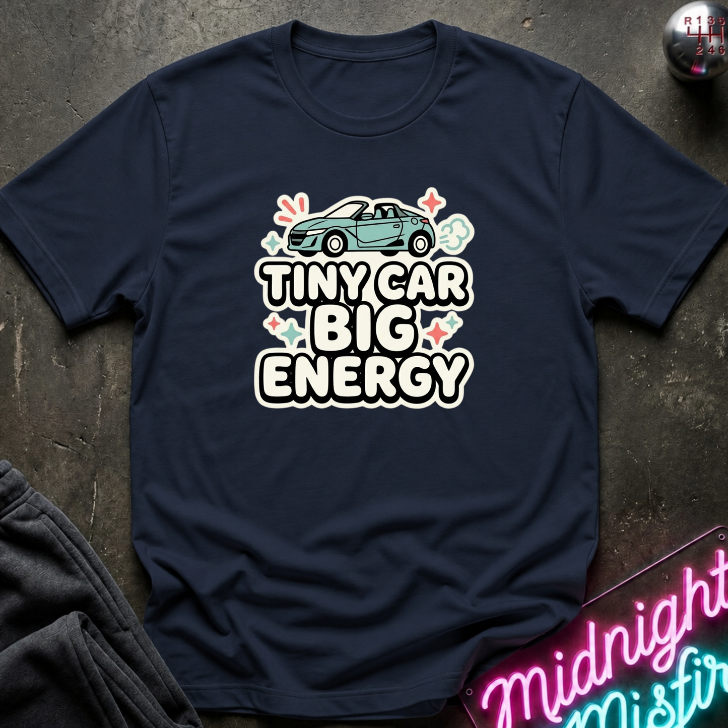 A navy blue short-sleeve t-shirt laid flat on a dark textured surface, featuring a graphic print of a small teal convertible car above the words 'TINY CAR BIG ENERGY' in white outlined letters with pastel accents.