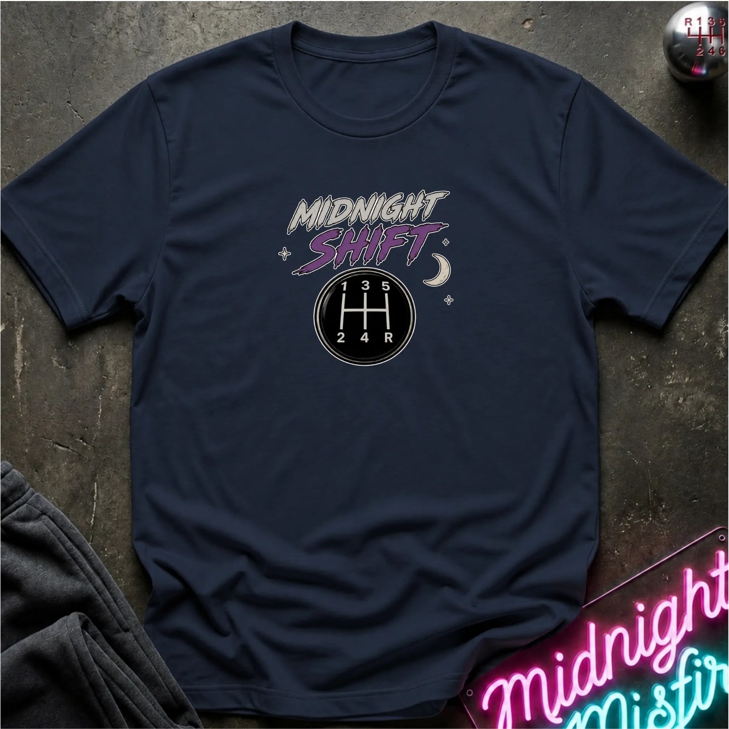 A navy blue short-sleeve t-shirt laid flat on a dark textured surface, featuring a 'Midnight Shift' graphic with a 5-speed manual gearshift pattern and stars. A neon sign and folded dark clothing are partially visible in the background.