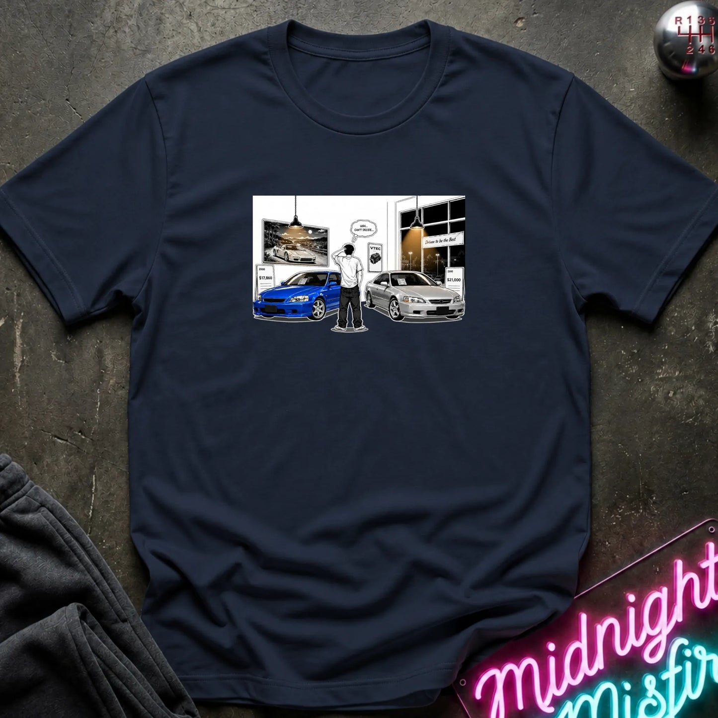 A navy blue short-sleeve t-shirt laid flat on a dark textured surface, featuring a rectangular graphic print on the chest with a blue car, a white car, and a small illustrated figure. 
