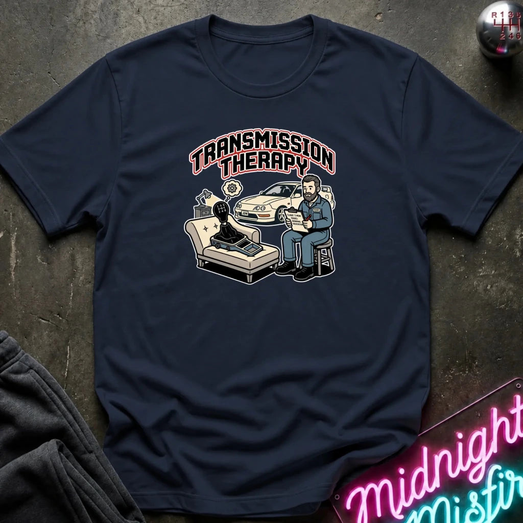 A navy blue graphic t-shirt laid flat on a dark textured surface. The shirt features a retro-style illustration of a therapist sitting next to a patient on a chaise lounge with a police car, under the text 'TRANSMISSION THERAPY'.