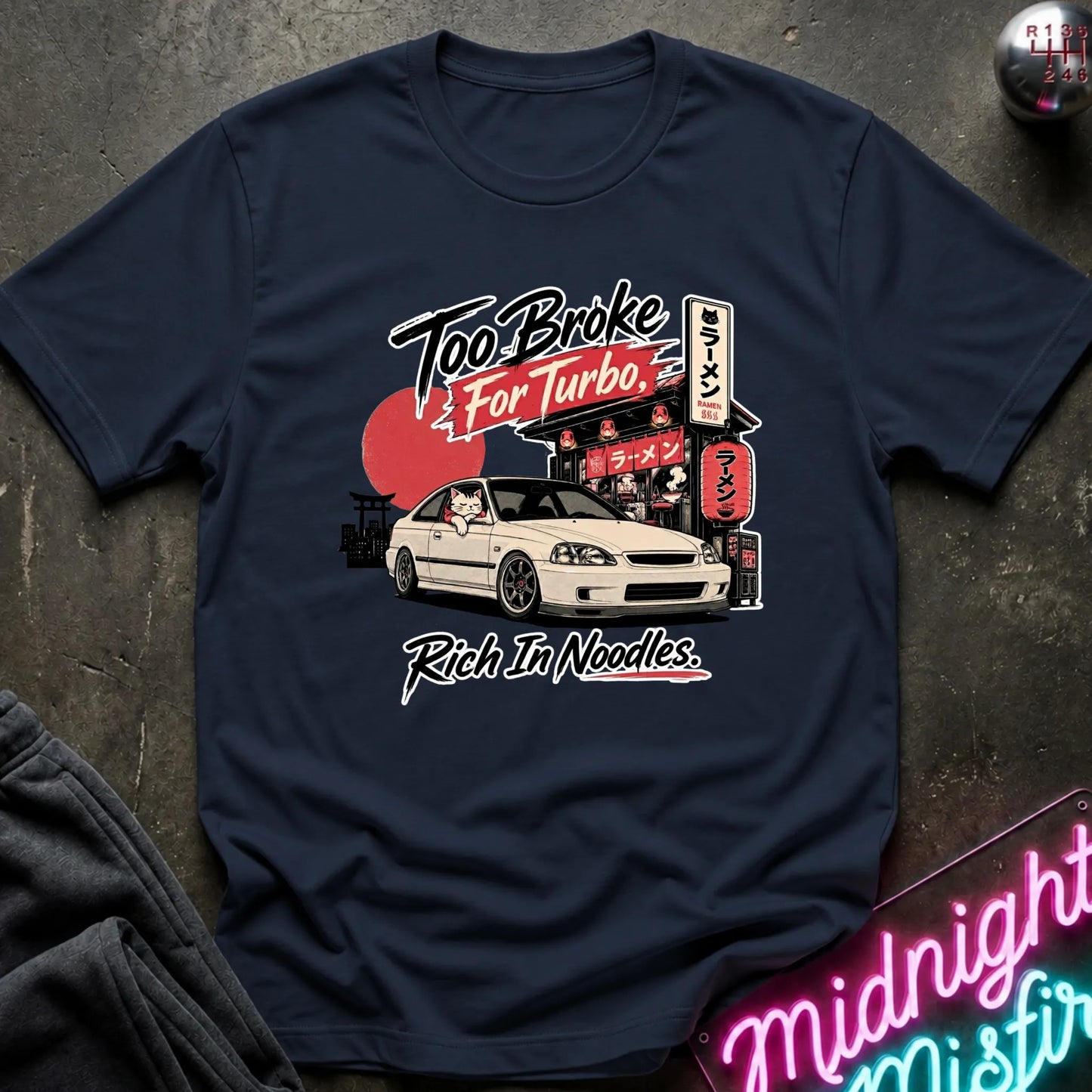 Navy short-sleeve graphic T-shirt laid flat on a dark textured surface. The shirt features a printed design of a white sports car in front of a Japanese-style street scene with a red sun and neon signs, plus the text 'Too Broke For Turbo' and 'Rich In Noodles.