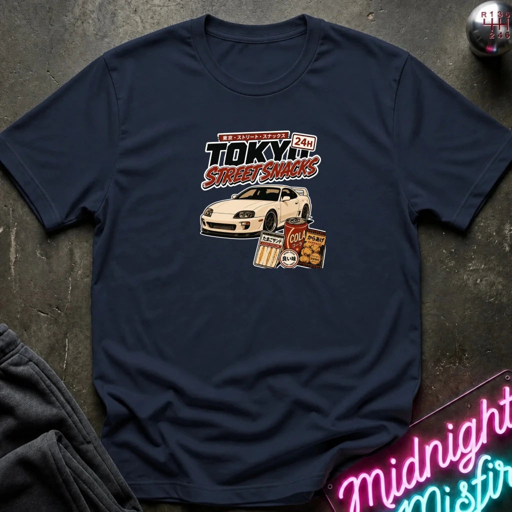 navy short-sleeve graphic T-shirt laid flat on a dark textured surface. The shirt features a central graphic with the text 'Tokyo Street Snacks' above a white sports car and snack illustrations.