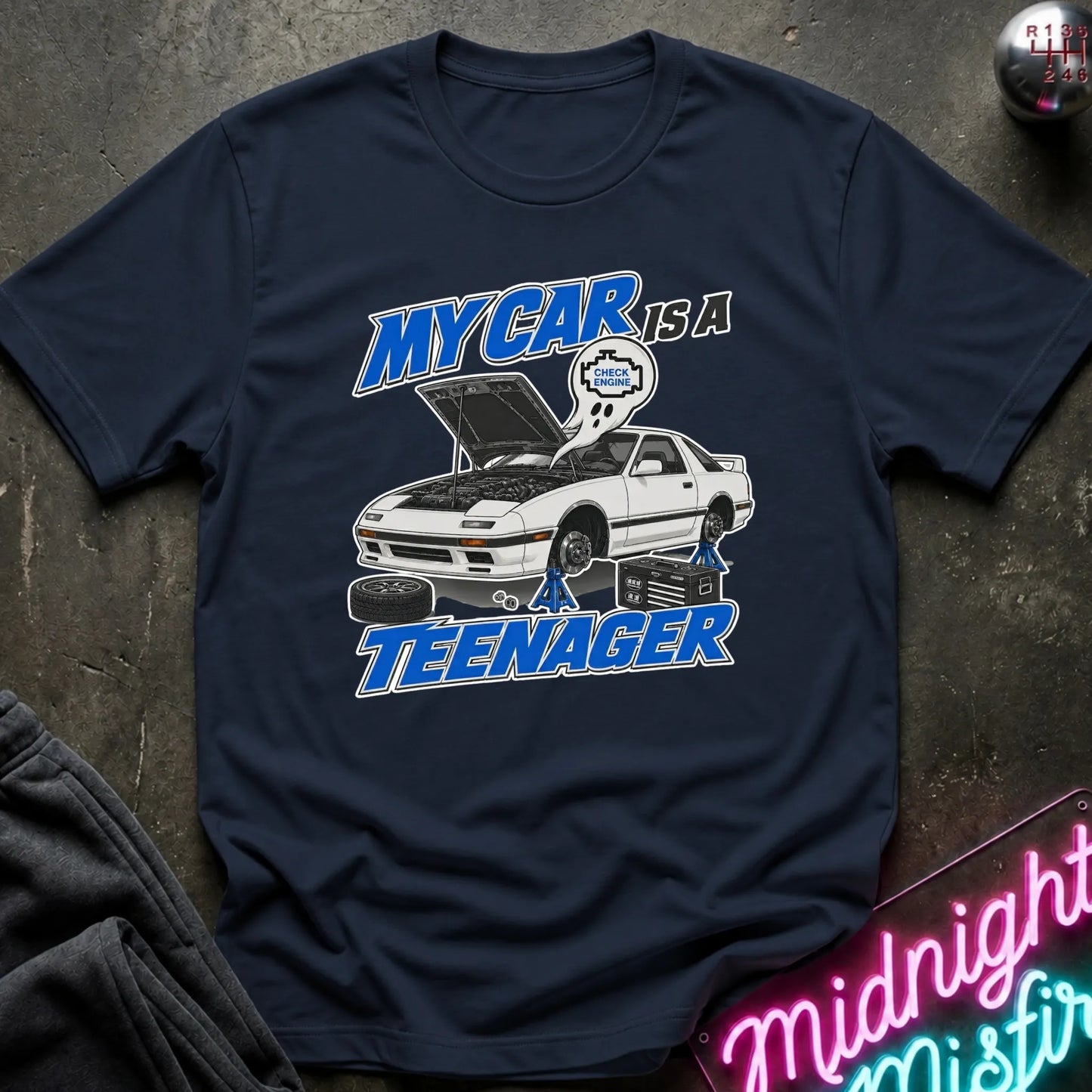 A Navy short-sleeve graphic T-shirt laid flat on a dark textured surface. The shirt features a white car with its hood open, blue and white text reading 'MY CAR IS A TEENAGER,'.