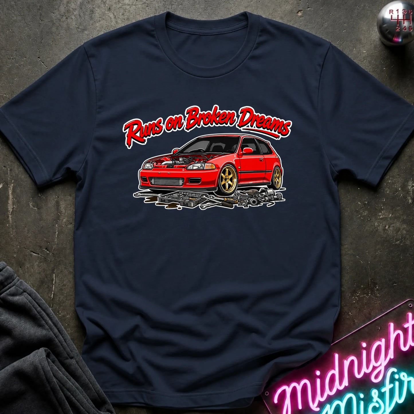 A navy blue graphic t-shirt laid flat on a dark textured surface. The shirt features a red sports car with gold wheels and the text 'Runs on Broken Dreams' in red and white.