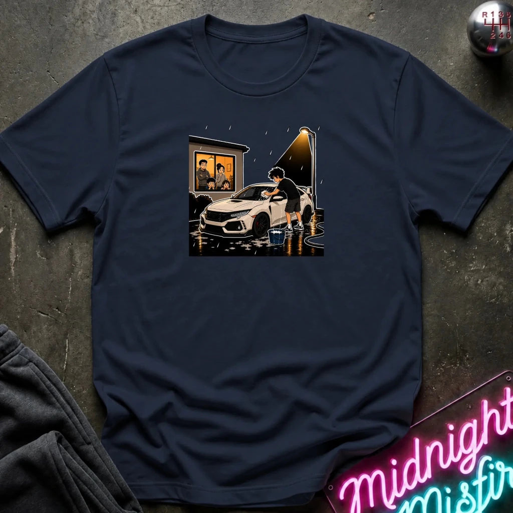 A navy blue short-sleeve t-shirt laid flat on a dark textured surface, featuring a central graphic print of a white car in a rainy street scene with a person and a building