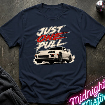 A navy blue graphic t-shirt laid flat on a dark textured surface. The shirt features a white sports car and the text 'JUST ONE PULL' in white and red.