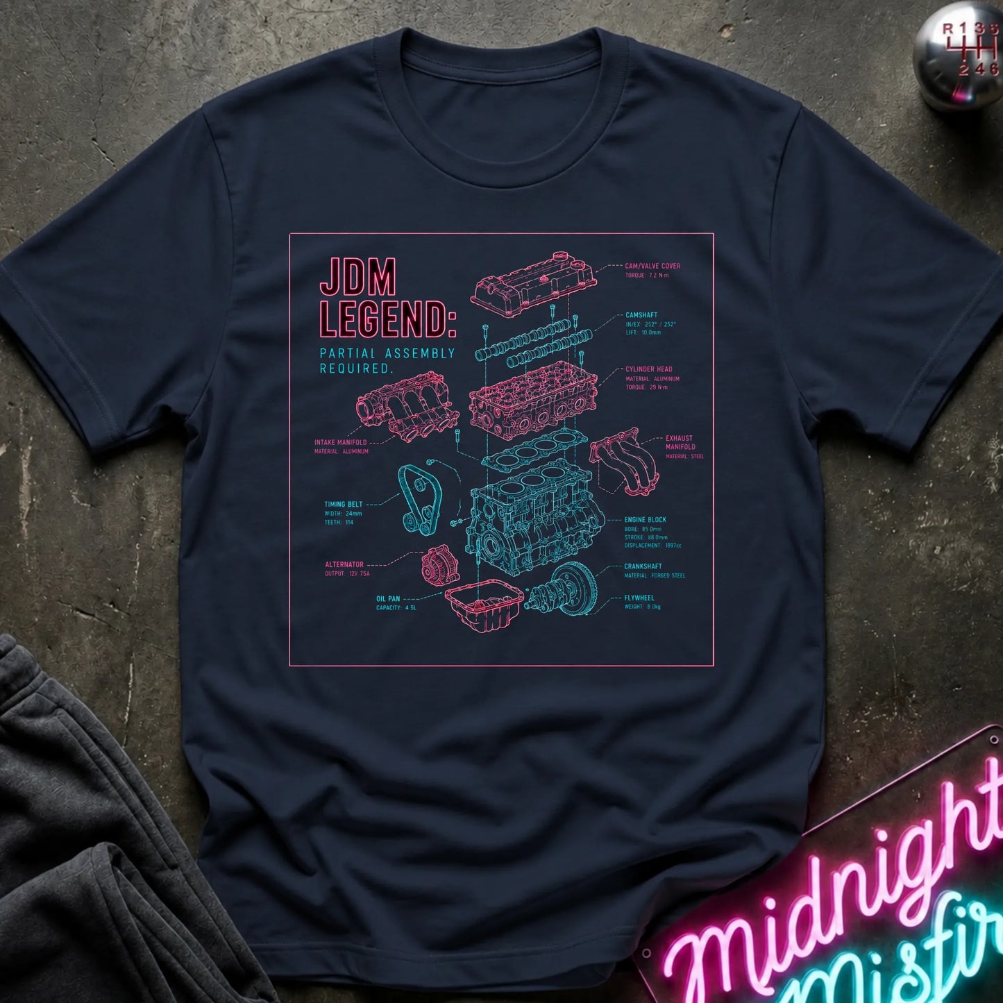 A navy blue graphic t-shirt laid flat on a dark textured surface. The shirt features a detailed pink and cyan exploded-view schematic of a car engine.