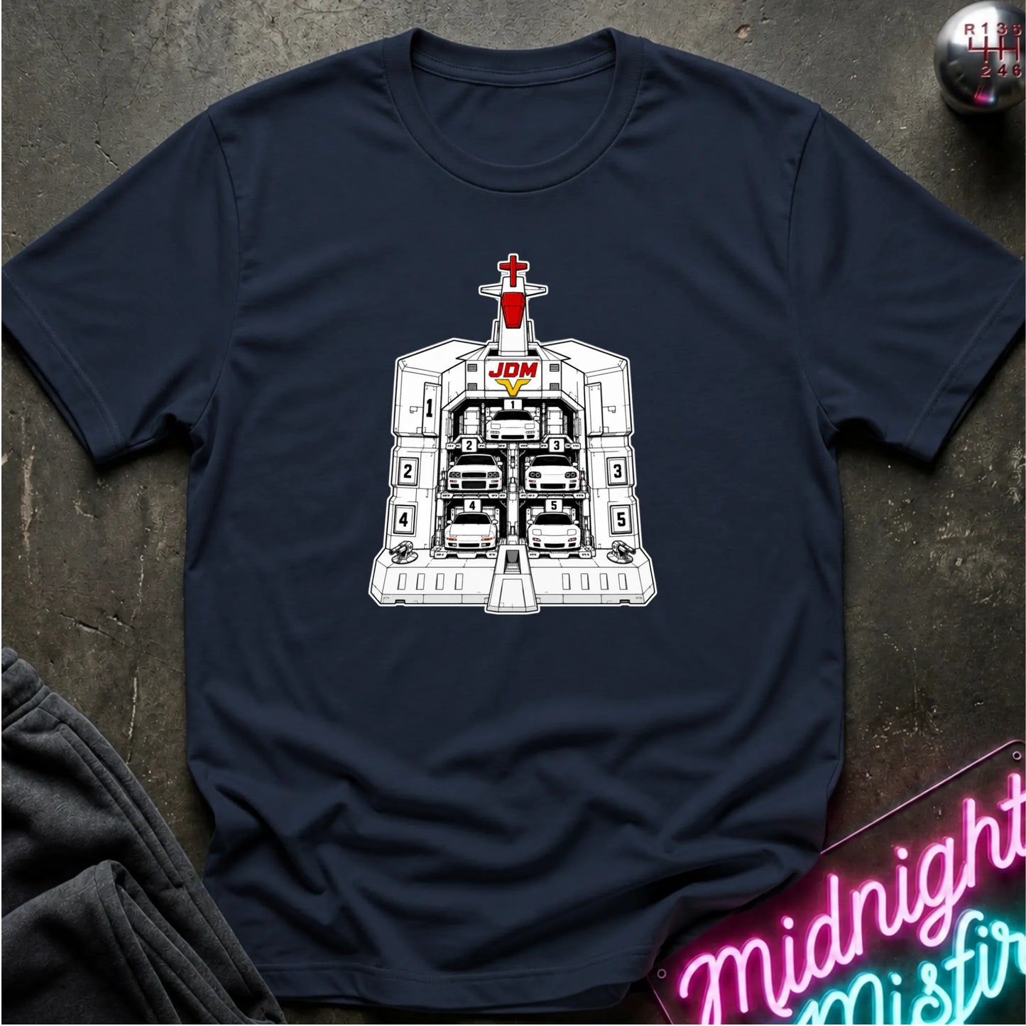 A navy blue short-sleeve crewneck t-shirt laid flat on a dark textured surface, featuring a black and white graphic of the JDM (Japanese Domestic Market) cars in a launch bay.