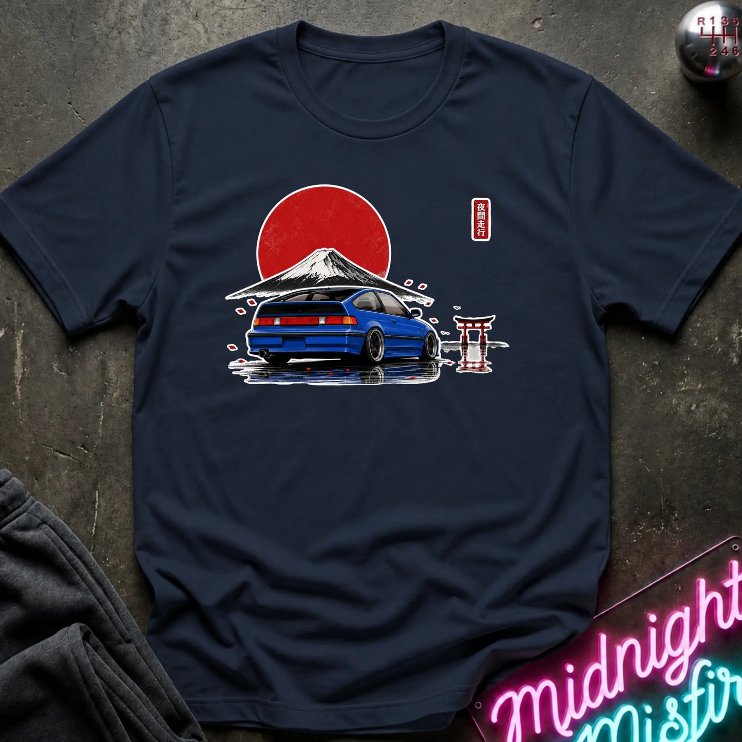 A navy blue graphic t-shirt laid flat on a dark textured surface. The graphic features a blue sports car in front of a red sun and Mount Fuji, with a small red and white torii gate and a vertical red label.