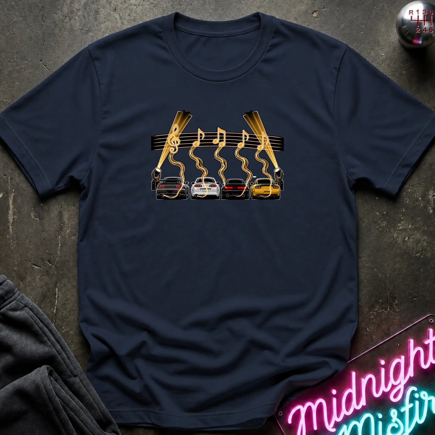 A navy blue short-sleeve t-shirt laid flat on a dark textured surface, featuring a graphic print of four cars (red, white, gray, yellow) with gold wavy lines and musical notes above them, flanked by two  lights.