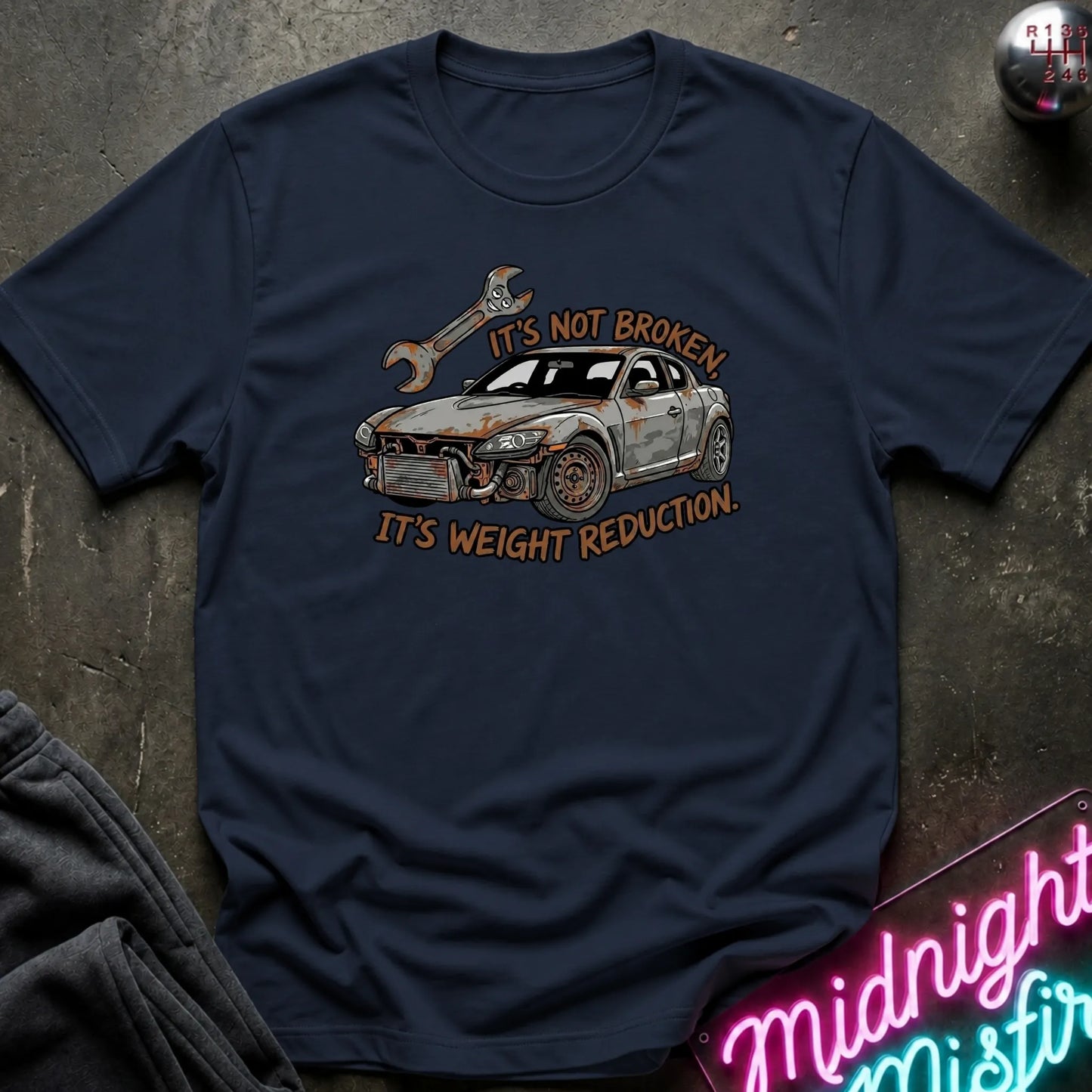 A navy blue graphic t-shirt laid flat on a dark textured surface. The shirt features a distressed graphic of a white and gray sports car with a wrench, accompanied by the text 'IT'S NOT BROKEN.