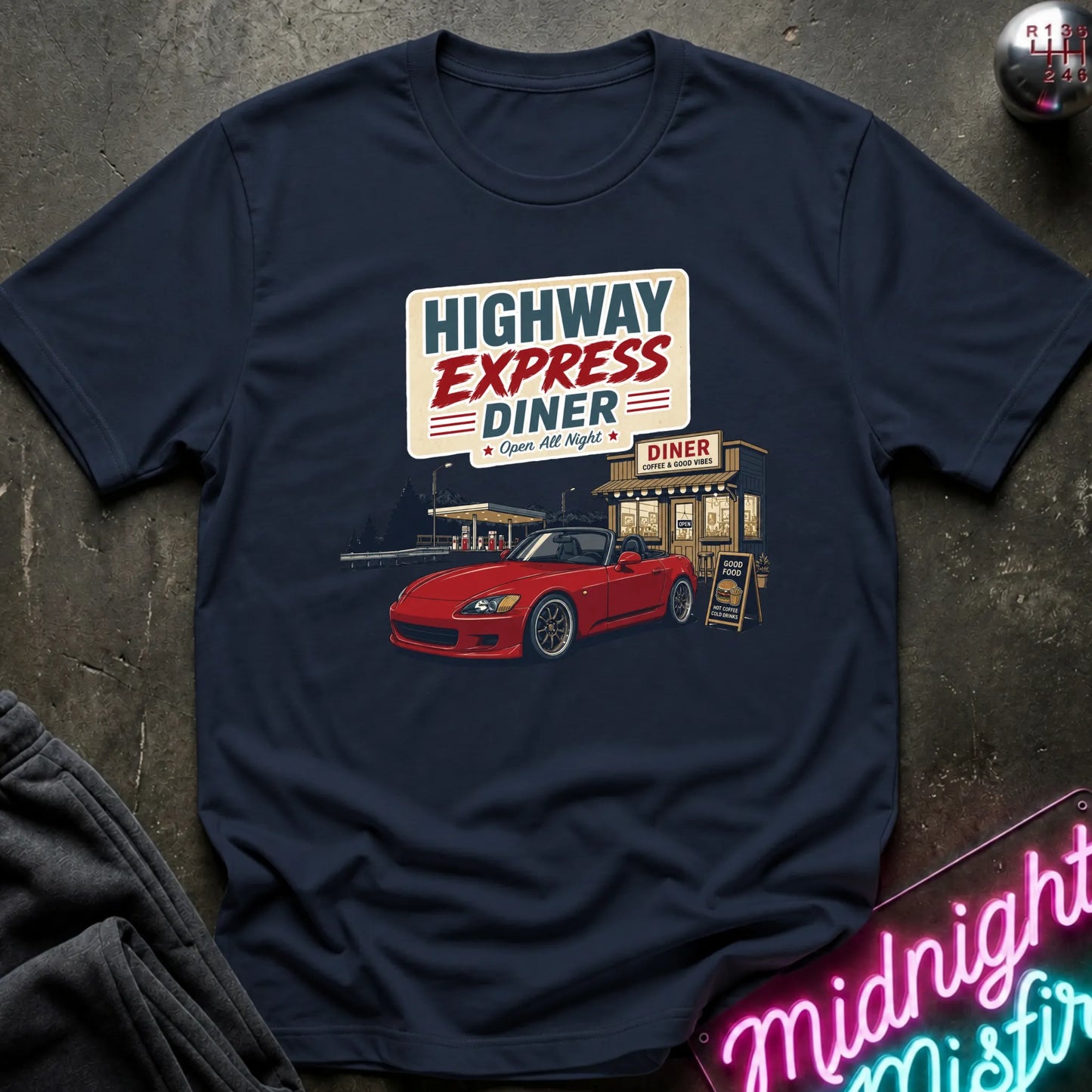 A navy blue short-sleeve t-shirt with a retro graphic of a red sports car at a diner under a sign reading 'HIGHWAY EXPRESS DINER', laid flat on a dark textured surface with a neon sign and folded fabric visible at the edges.