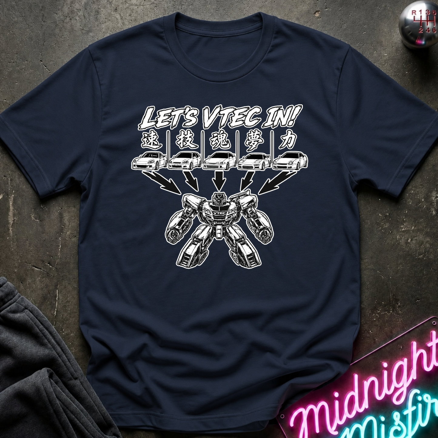 A navy blue short-sleeve t-shirt with a white graphic print that reads 'LET'S VTEC IN!' above five cars transforming into an robot, laid flat on a dark textured surface with a neon sign partially visible in the corner.