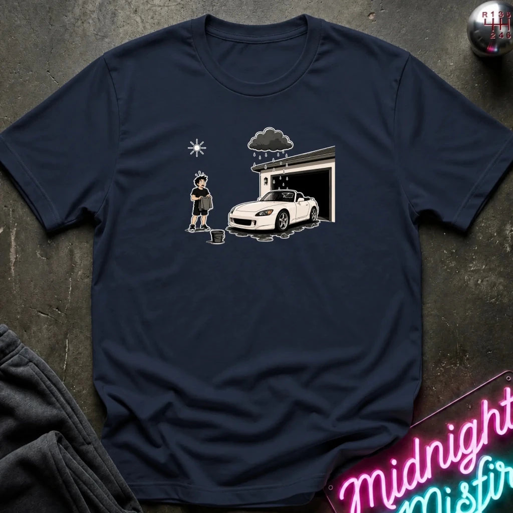 A navy blue short-sleeve t-shirt with a cartoon graphic of a person washing a white sports car in front of a garage under a dark raincloud, laid flat on a dark textured surface.