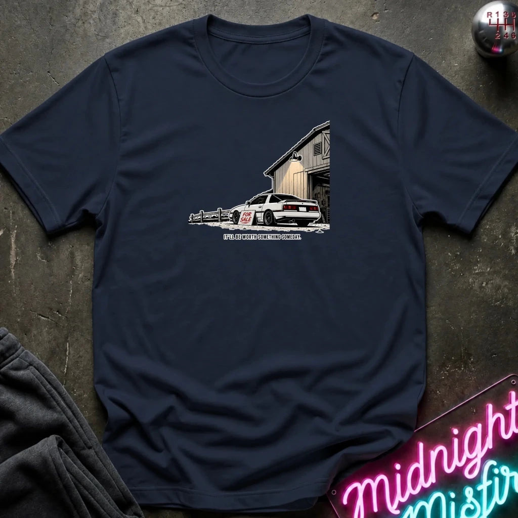 A navy blue short-sleeve t-shirt laid flat on a dark textured surface, featuring a graphic print of a white sports car parked in front of a barn 