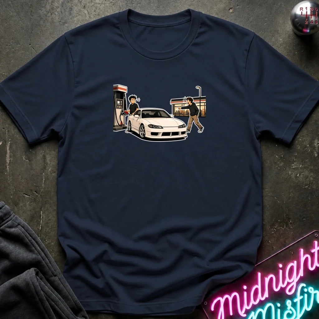 A navy blue short-sleeve t-shirt with a graphic print of a white sports car at a gas station, laid flat on a dark textured surface with a neon sign and folded sweatpants nearby.