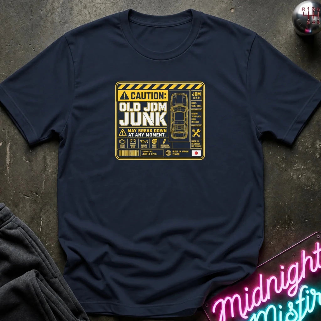 A navy blue  short-sleeve t-shirt laid flat on a dark textured surface, featuring a yellow and black caution-style graphic on the chest that reads 'OLD JDM JUNK' with a car illustration and warning symbols.