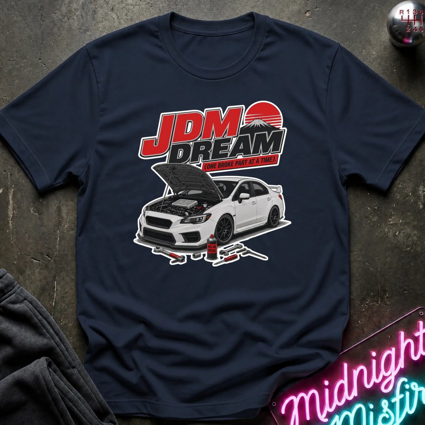 A navy blue graphic t-shirt laid flat on a dark textured surface. The shirt features a graphic of a white sports car with its hood open, beneath the red and white text 'JDM DREAM' and a red sun graphic.