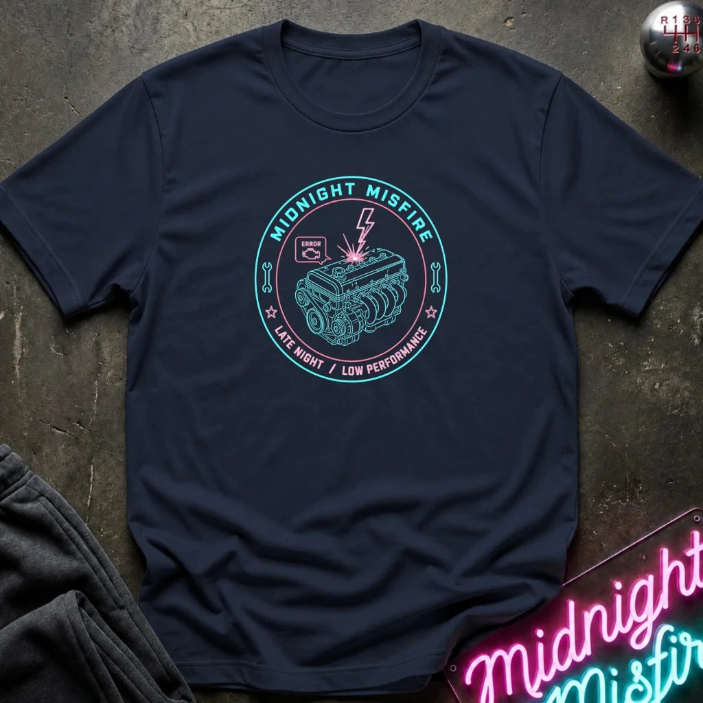 A navy blue crewneck t-shirt laid flat on a dark textured surface. The center of the shirt features a circular graphic with a teal engine illustration, a lightning bolt, and the words 'MIDNIGHT MISFIRE' and 'LATE NIGHT / LOW PERFORMANCE'.