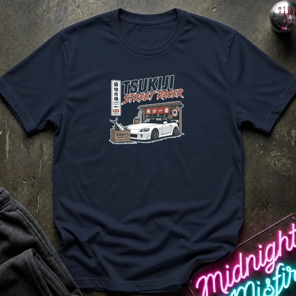 A navy blue crewneck t-shirt laid flat on a dark textured surface, featuring a graphic print of a white sports car and a street racer shop with the text 'TSUKIJI STREET RACER'.
