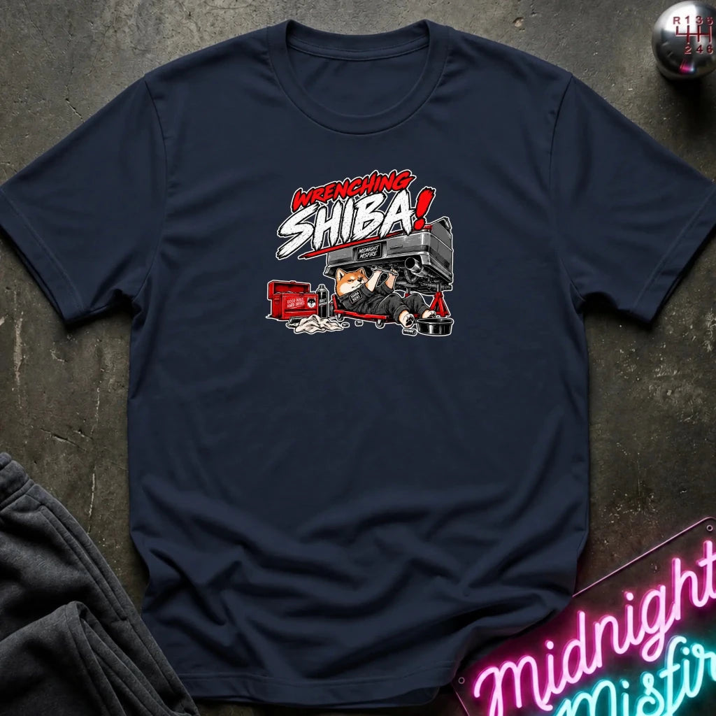 A navy blue short-sleeve graphic t-shirt laid flat on a dark textured surface. The graphic features the text 'WRENCHING SHIBA!' in red and white comic-style lettering above an illustration of a character in a black and red vehicle.