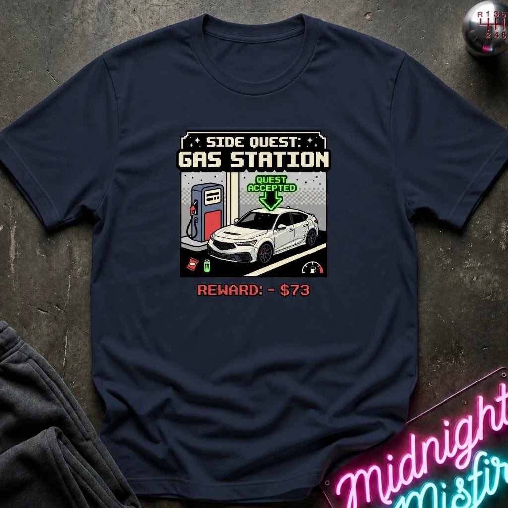 A navy blue graphic t-shirt laid flat on a dark textured surface. The shirt features a retro video game style 'Side Quest: Gas Station' graphic with a white car, a gas pump, and a green 'Quest Accepted' arrow.