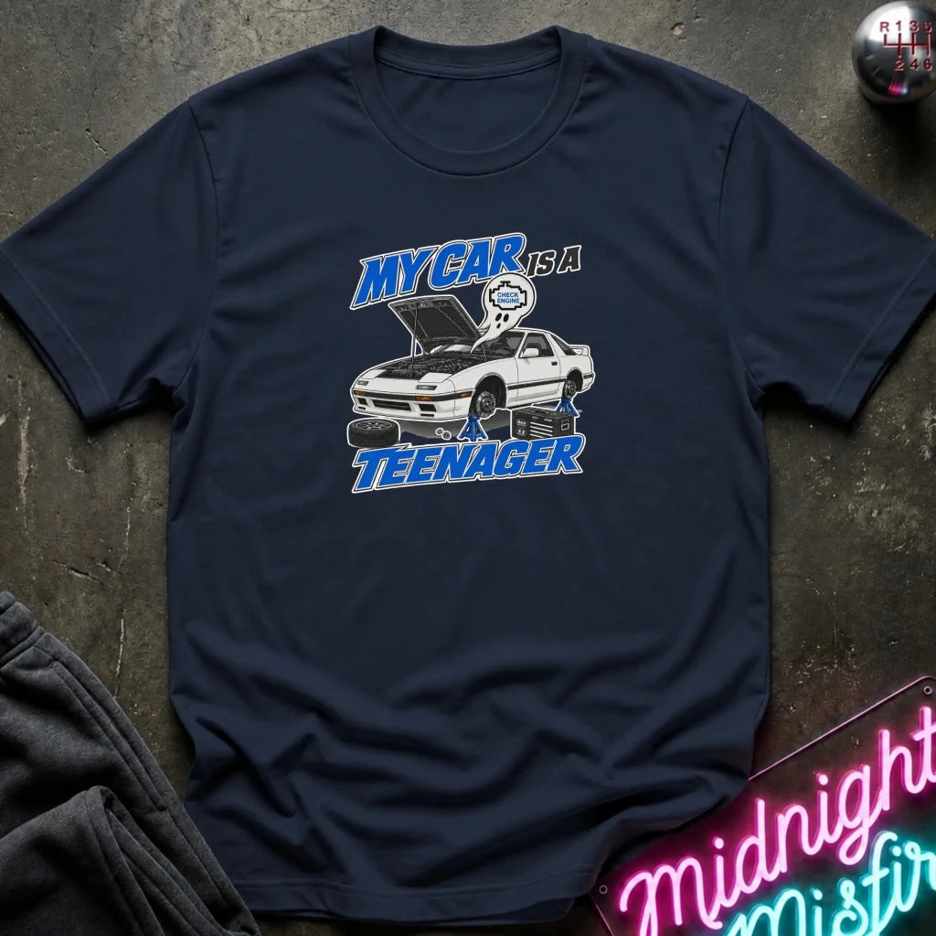 A navy blue graphic t-shirt laid flat on a dark textured surface. The shirt features a white and blue graphic of a white sports car with its hood open, accompanied by the text 'MY CAR IS A TEENAGER' in blue and white.