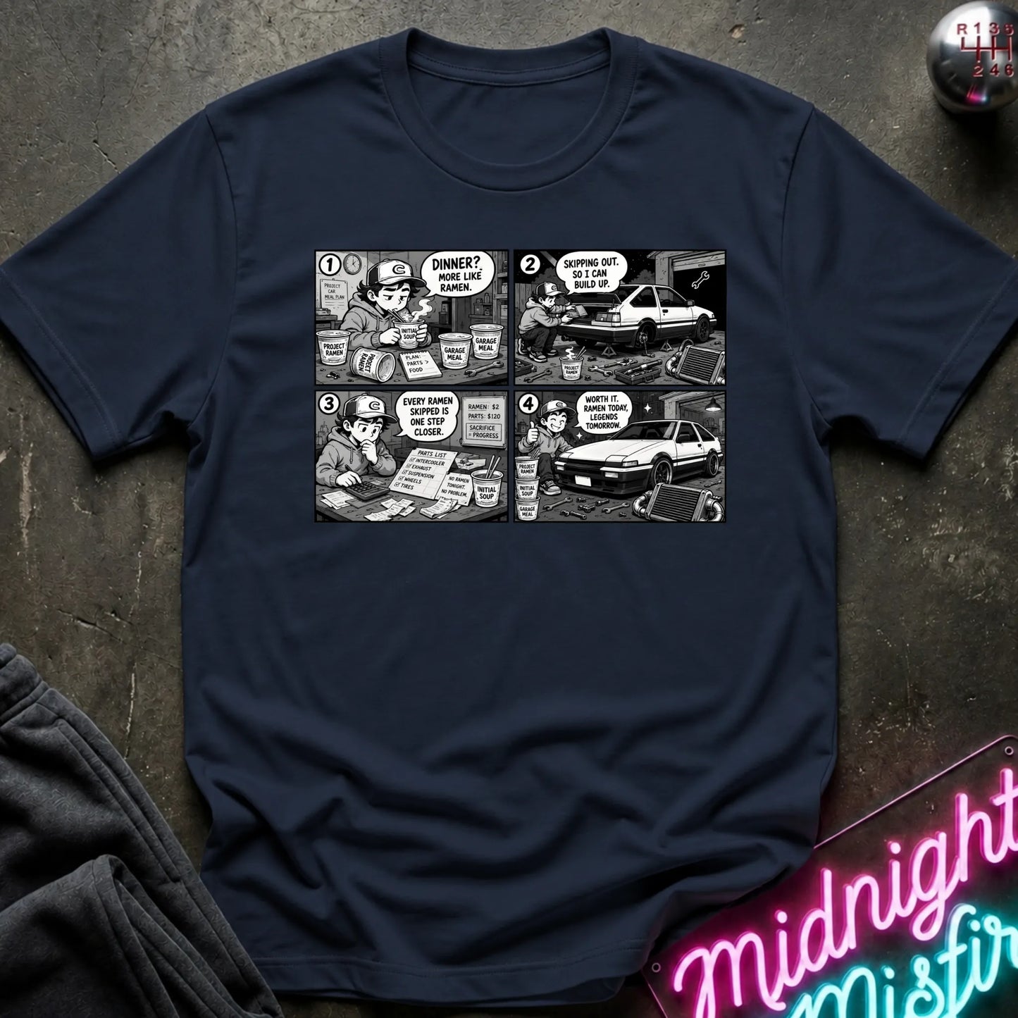 A navy blue short-sleeve crewneck t-shirt featuring a black and white comic strip graphic on the chest, styled in a flat-lay on a dark textured surface with a neon sign and dark pants partially visible.