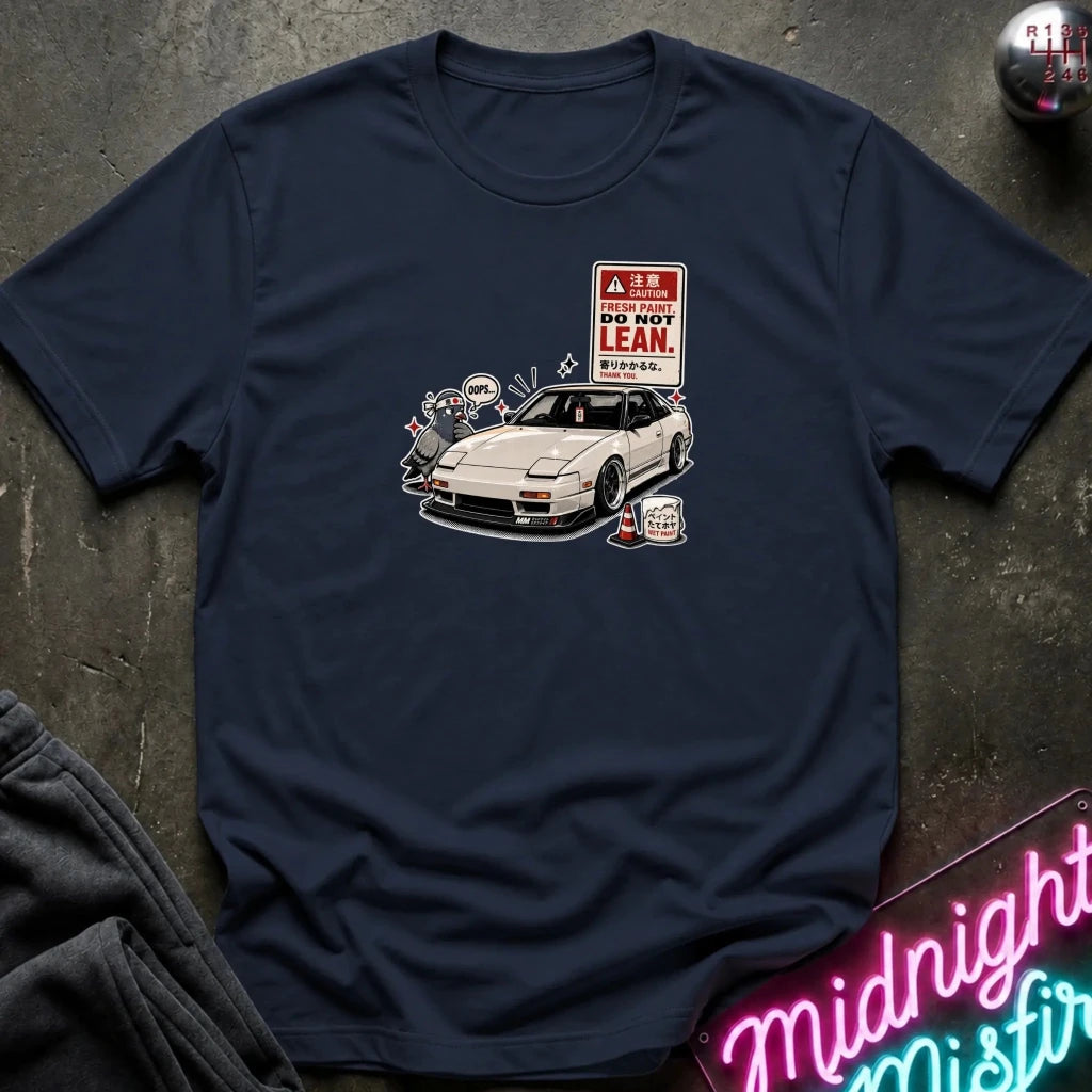 A navy short-sleeve t-shirt laid flat on a dark textured surface, featuring a graphic print of a white sports car next to a 'DO NOT LEAN' warning sign.