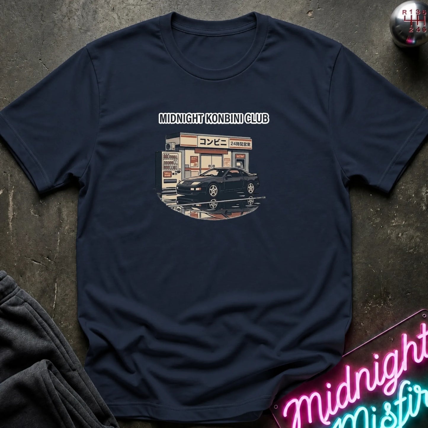 A navy blue short-sleeve t-shirt with a vintage-style graphic of a black sports car parked outside a convenience store labeled 'MIDNIGHT KONBINI CLUB'. The shirt is laid flat on a dark textured surface with a neon sign and dark pants partially visible in the corners.