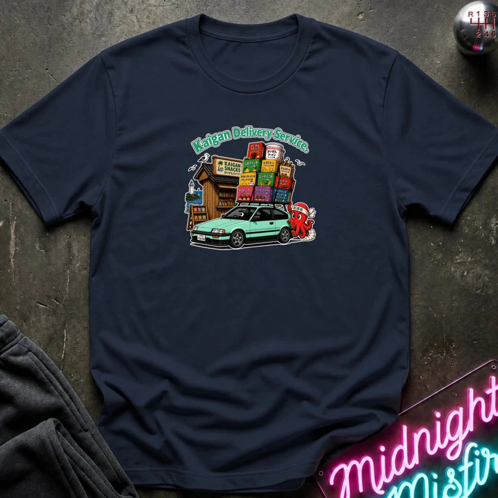A navy blue graphic t-shirt laid flat on a dark textured surface. The graphic features a light green car, and a small building with colorful signs under the text 'Kaigan Delivery Service'.