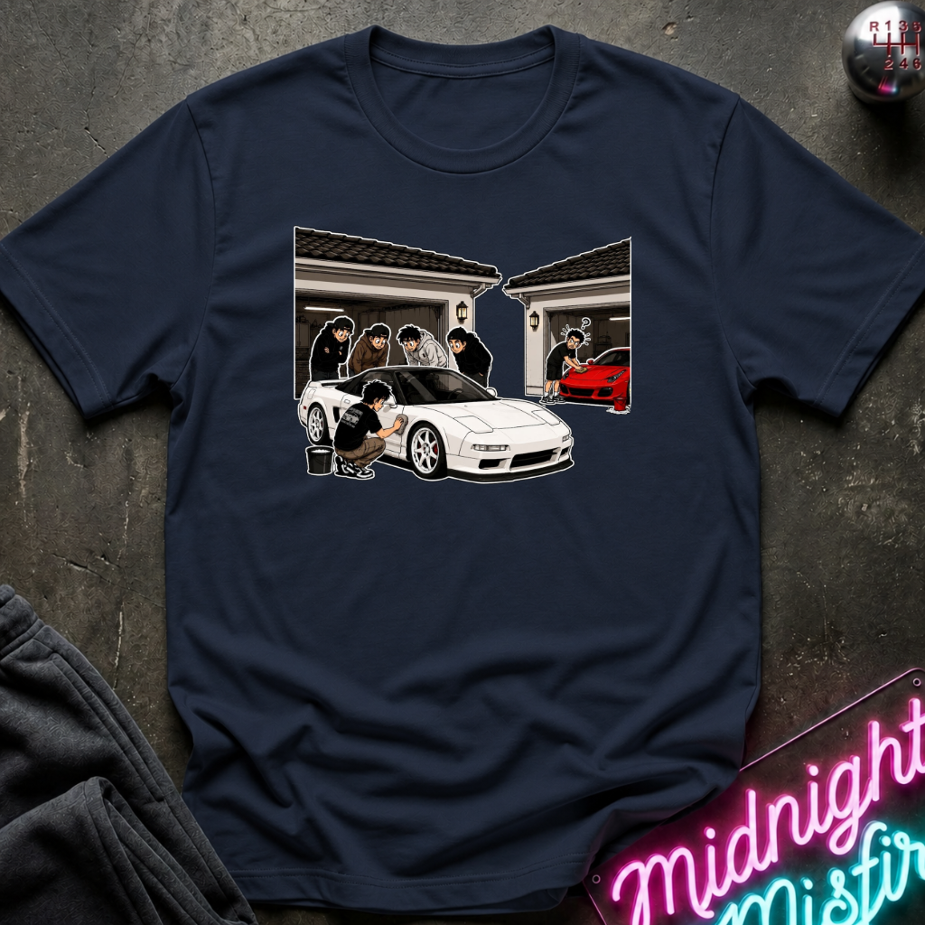 A navy blue short-sleeve t-shirt with a graphic print of a white sports car and people in front of garages, laid flat on a dark textured surface with a neon sign and sweatpants partially visible.