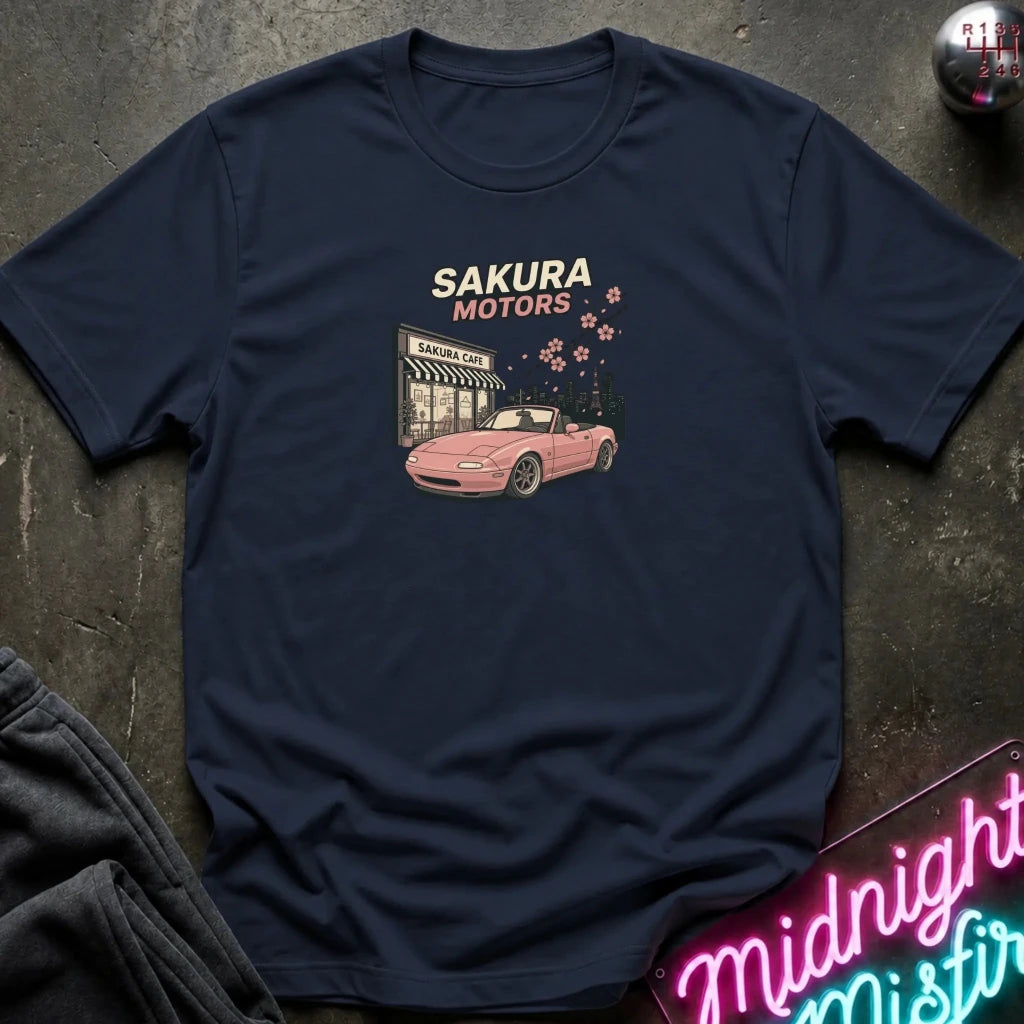 A navy blue graphic t-shirt laid flat on a dark textured surface. The shirt features a retro-style graphic of a pink convertible car in front of a 'Sakura Motors' shop, with cherry blossoms and the text 'SAKURA MOTORS' printed on the chest.