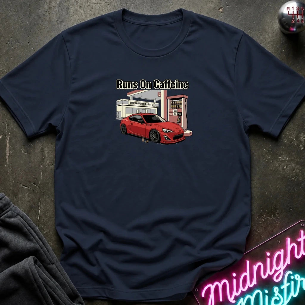 A navy blue short-sleeve t-shirt laid flat on a dark textured surface, featuring a graphic of a red sports car in front of a gas station with the text 'Runs On Caffeine'.