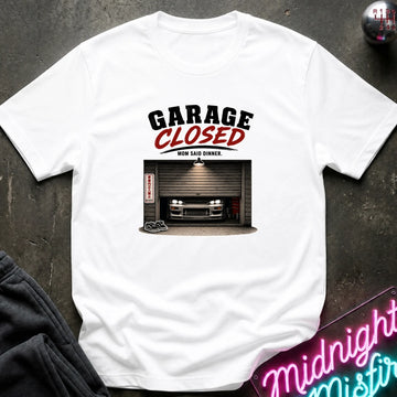A white short-sleeve graphic t-shirt laid flat on a dark textured surface. The graphic features the text 'GARAGE CLOSED' in black and red, above an illustration of a car parked inside a garage.
