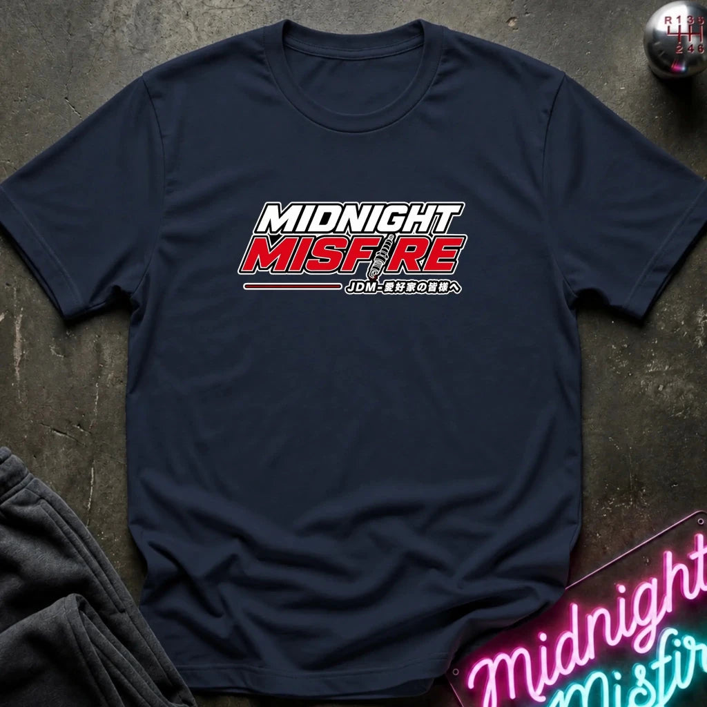 A navy blue short-sleeve t-shirt with a 'MIDNIGHT MISFIRE' graphic printed on the chest, laid flat on a dark textured surface with a neon sign and dark pants in the background.