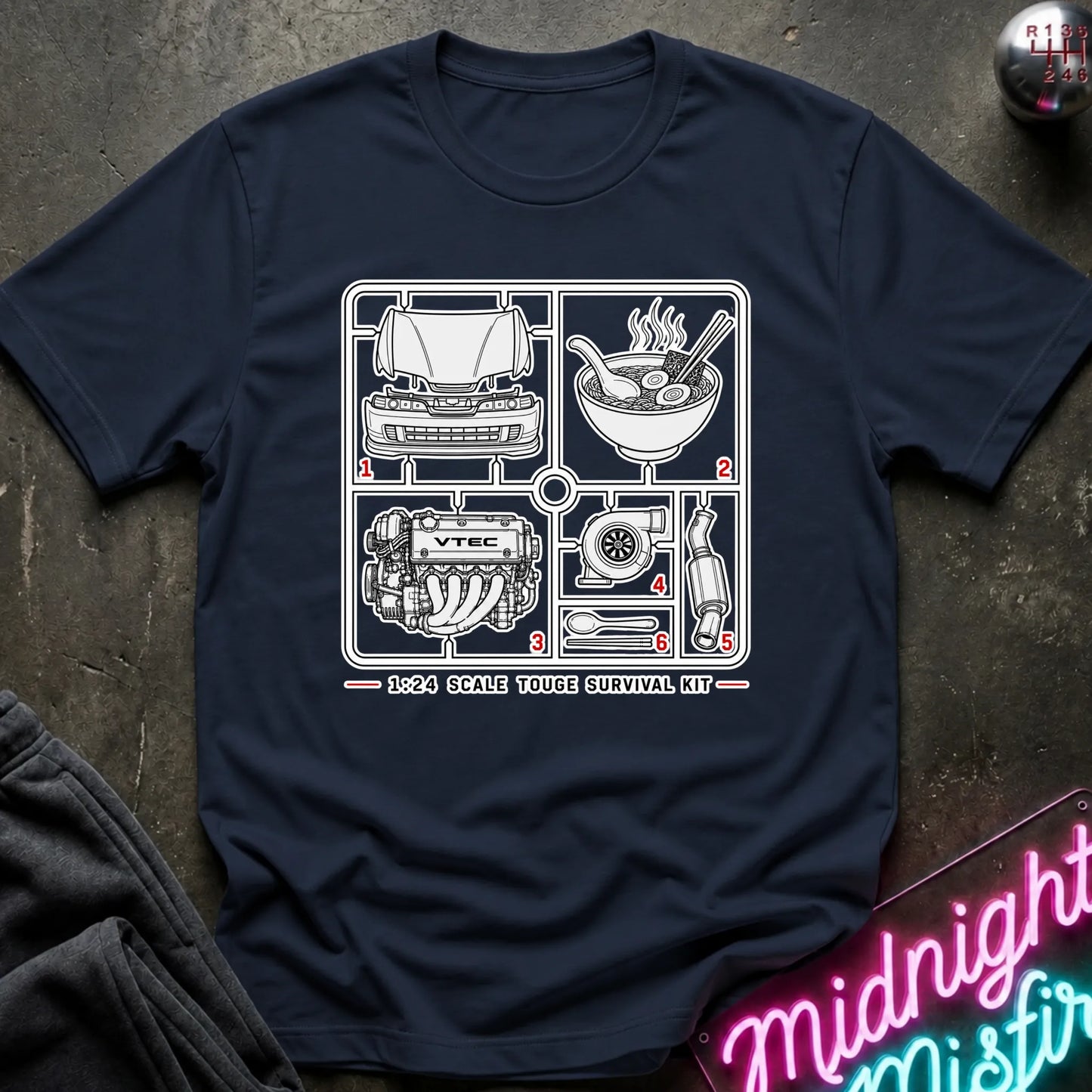 A navy blue short-sleeve graphic T-shirt laid flat on a dark textured surface. The shirt features a black line-art schematic of a survival kit with numbered illustrations and the text '1:24 SCALE TOUGHE SURVIVAL KIT' printed on the chest.