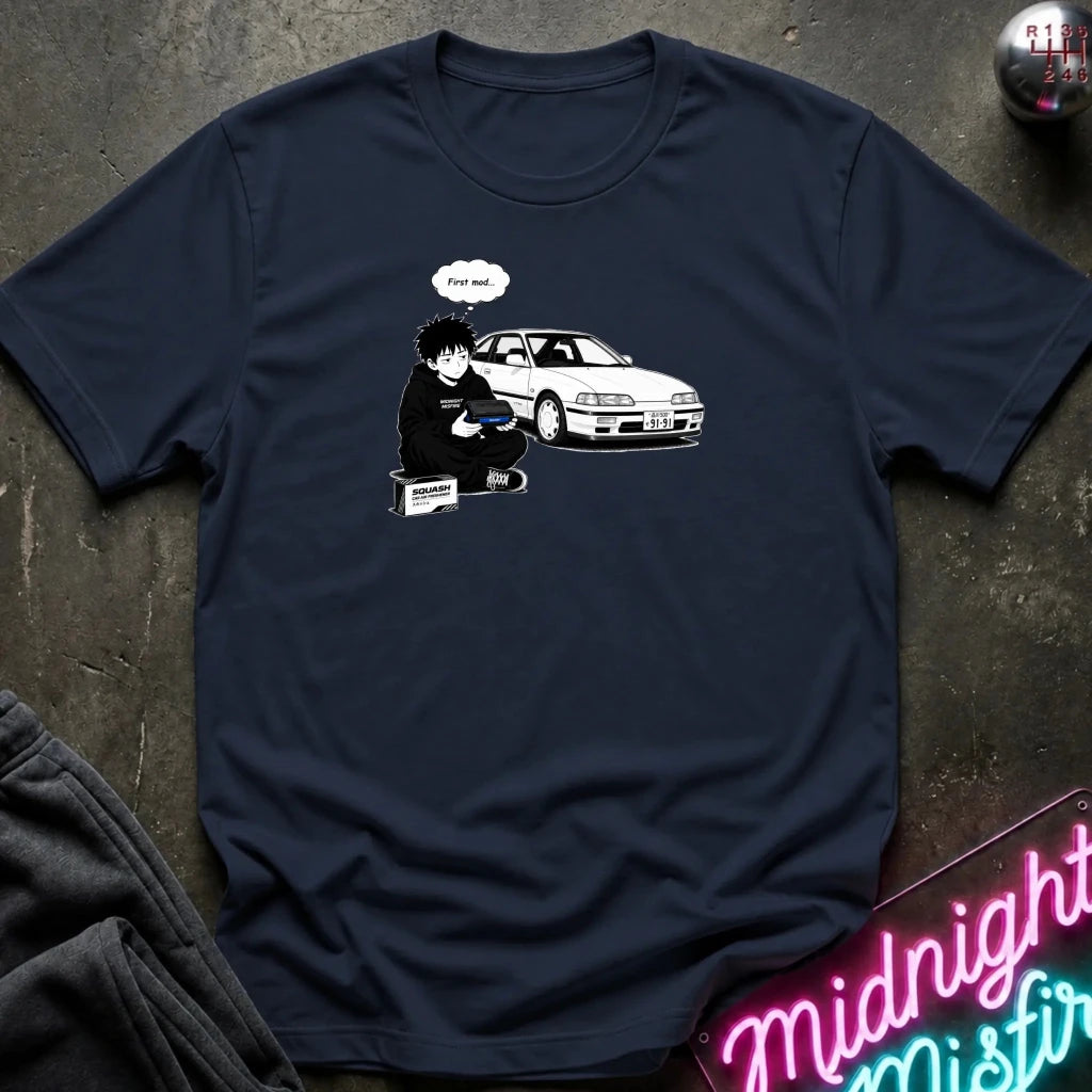 A navy blue short-sleeve t-shirt with a graphic print of a cartoon character next to a white car, laid flat on a dark textured surface with a neon sign and dark pants partially visible.