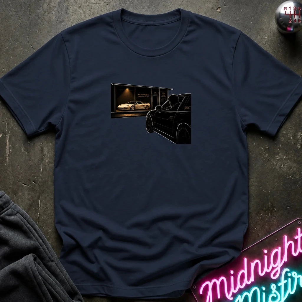 A navy blue crewneck t-shirt laid flat on a dark textured surface, featuring a graphic print of a black car in front of a lit garage. 
