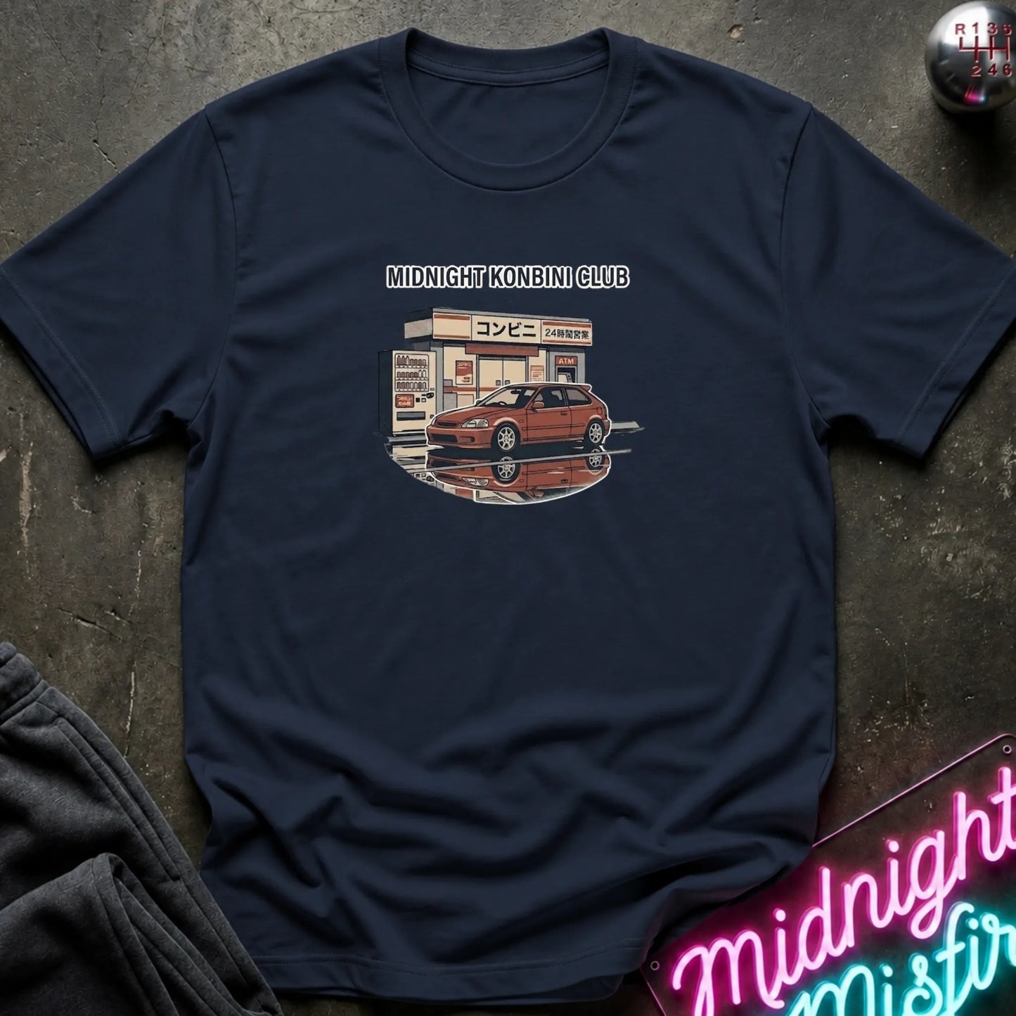 A navy blue short-sleeve t-shirt laid flat on a dark textured surface, featuring a graphic print of a red sports car parked in front of a convenience store with the text 'MIDNIGHT KONBINI CLUB' above it. A neon sign and dark clothing are partially visible as props around the shirt.