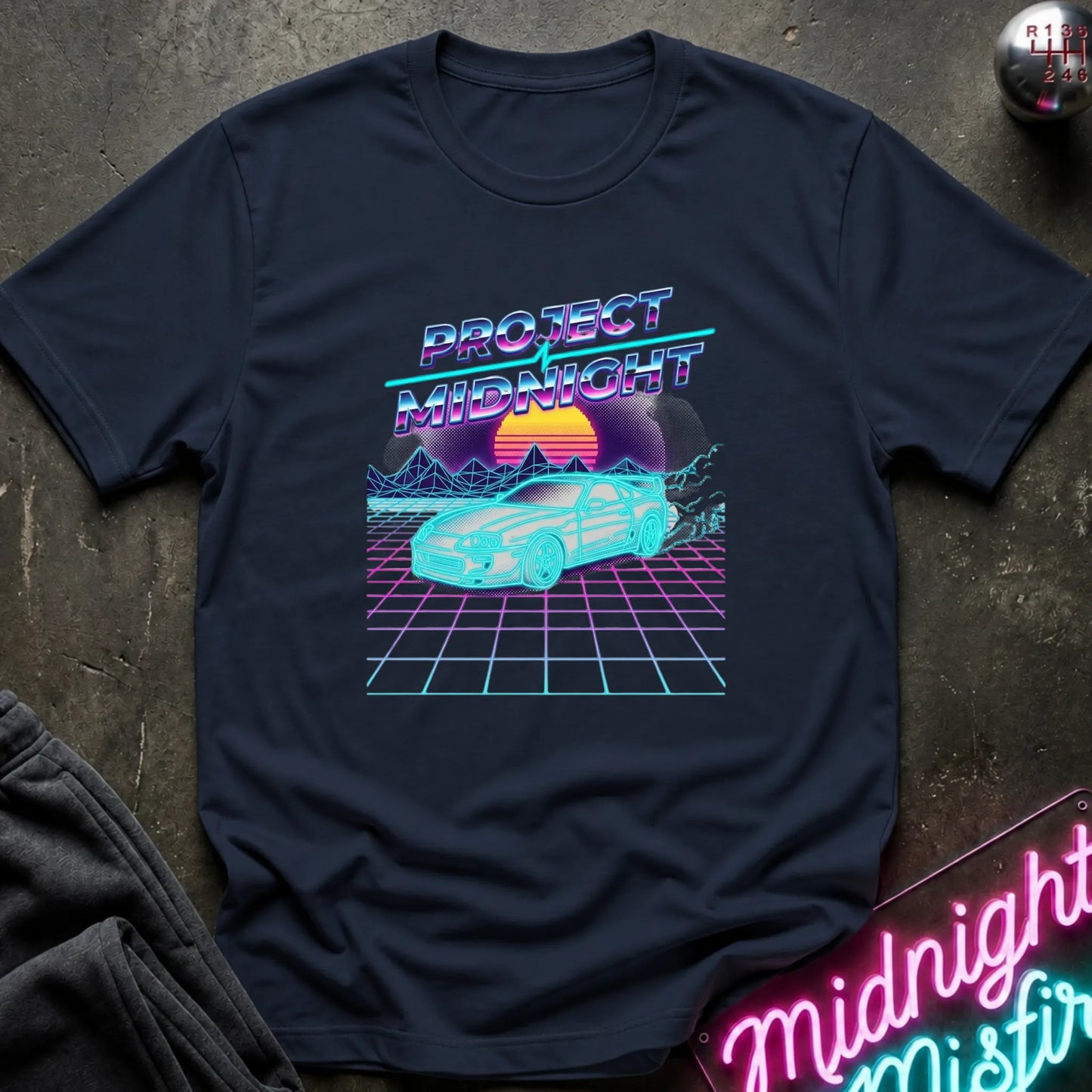 A navy blue graphic t-shirt laid flat on a dark textured surface. The shirt features a retro 80s synthwave graphic with the text 'PROJECT MIDNIGHT' in neon purple and blue, above a cyan car on a pink grid with a sunset.
