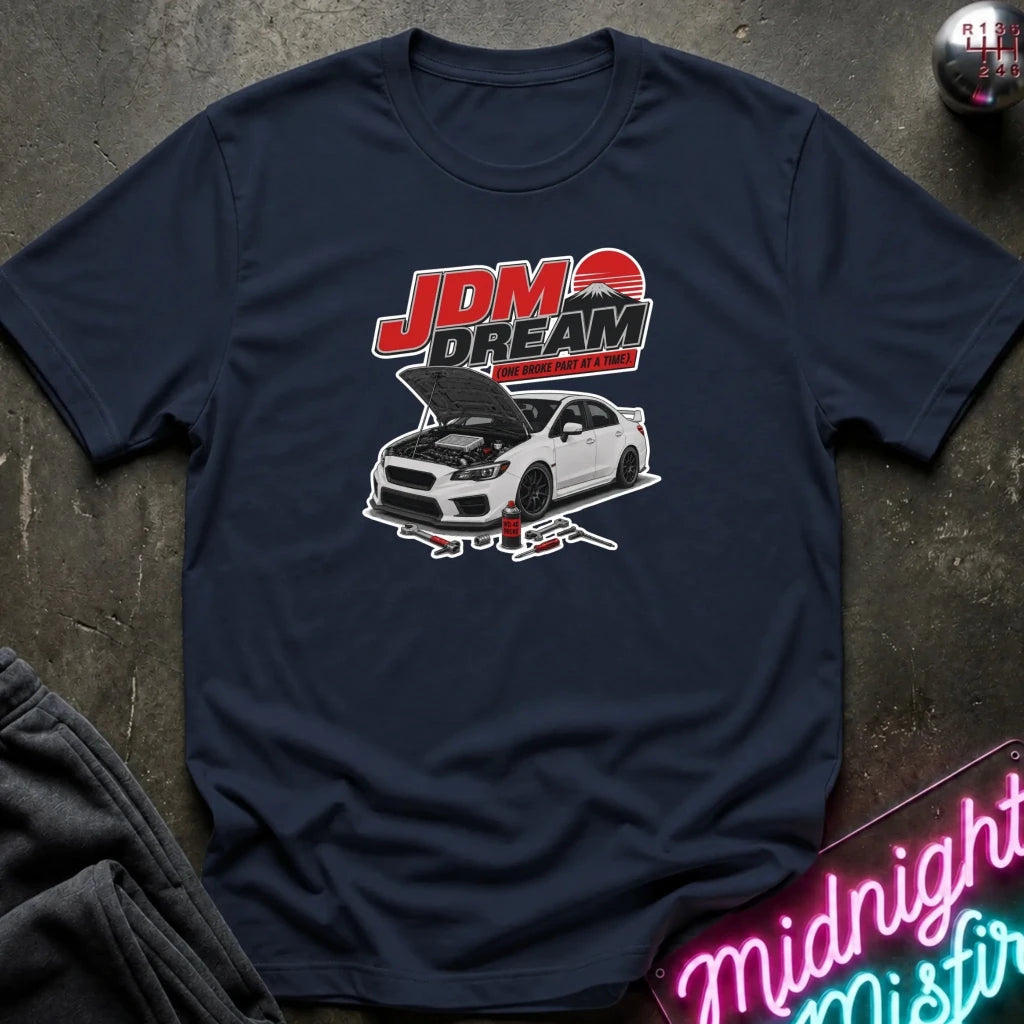 A navy blue graphic t-shirt laid flat on a dark textured surface. The shirt features a graphic print of a white sports car with its hood open, beneath the red and white text 'JDM DREAM'.
